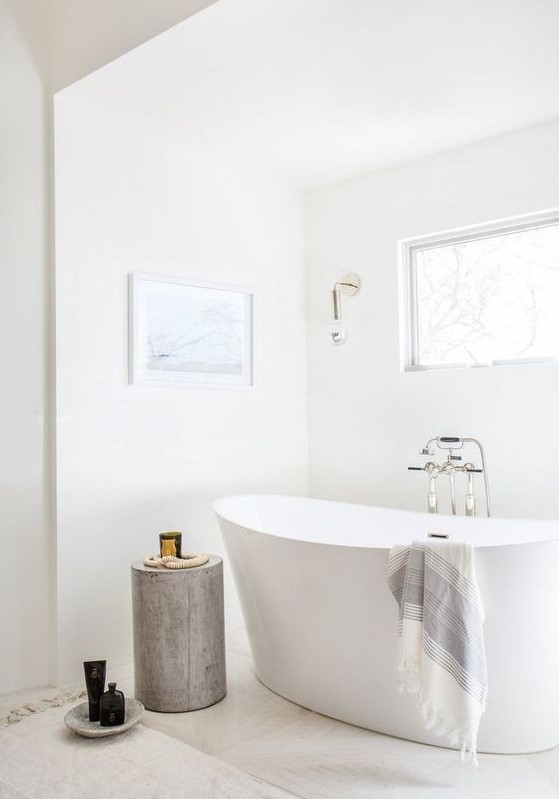 a serene white bathroom with marble tiles on the floor, a tub, a wooden stool, a window and some wall sconces