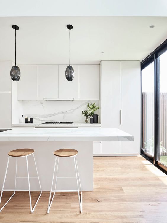 a sleek minimalist kitchen in white, with a white marble backsplash, black pendant lamps over the island and tall stools