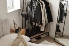 a small makeshift closet with a metal and wood rack for clothes and shoes and a floor mirror in a black frame