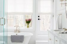 a traditional white bathroom with marble tiles clad in a chveron pattern, a tub by the window and a large vanity
