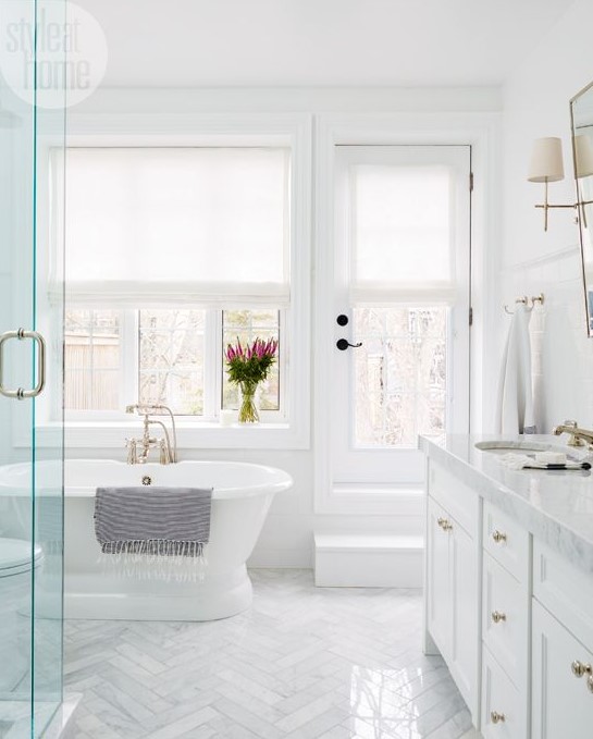 a traditional white bathroom with marble tiles clad in a chveron pattern, a tub by the window and a large vanity