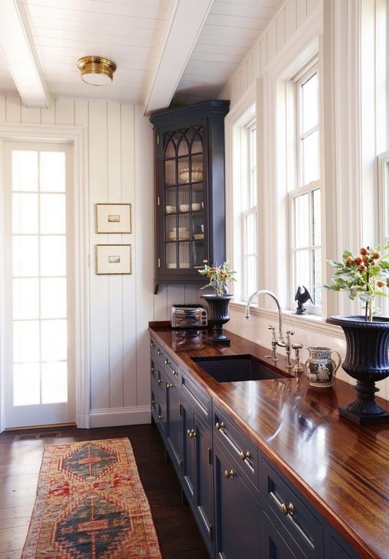 a vintage cottage kitchen with white beadboard walls, navy cabinets and a butcherblock countertop, a printed rug