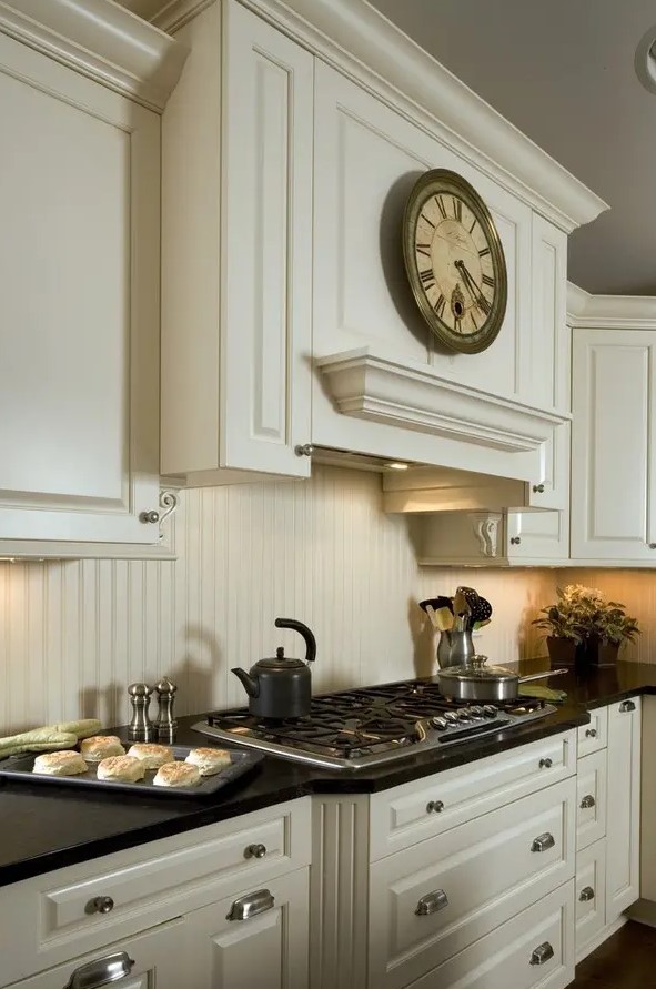 a vintage ivory kitchen with shaker style cabinets and a matching beadboard backsplash, black countertops and a clock is amazing