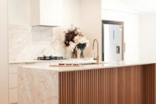 a warm neutral kitchen with white cabinets, a fluted kitchen island, pink marble countertops and a backsplash
