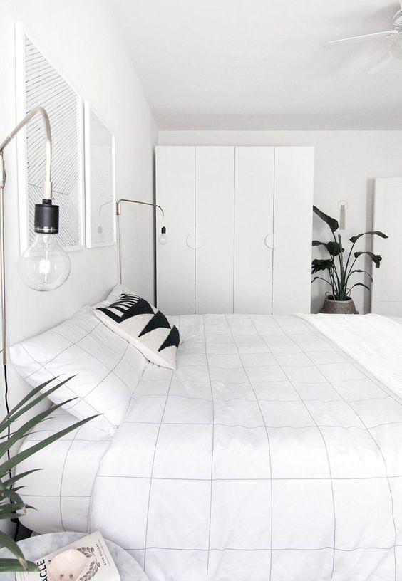 a white Scandinavian bedroom with an IKEA Pax wardrobe, a bed with graphic bedding, wall sconces and potted plants