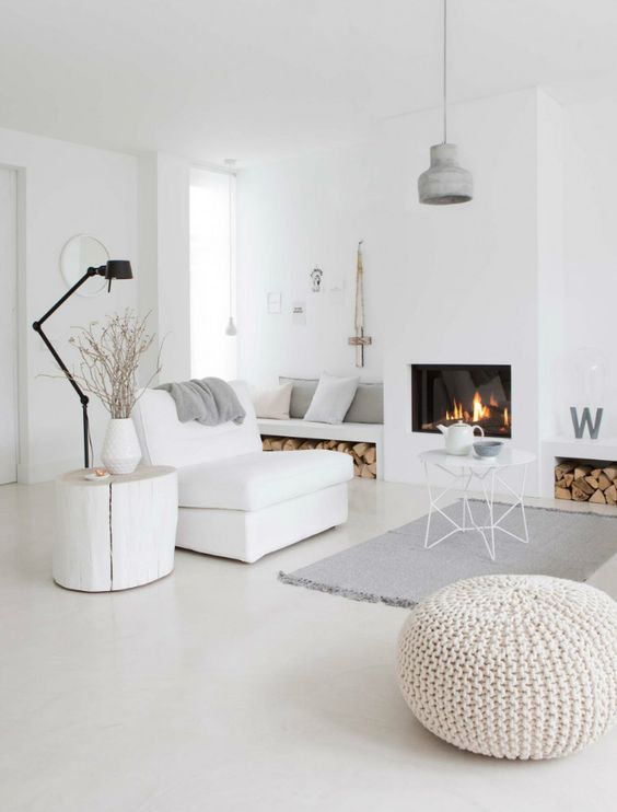 a white Scandinavian living room with built-in benches and firewood storage, a fireplace, a white chair, a table and some lamps