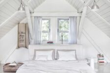 a white attic bedroom with an upholstered bed and white bedding, grey curtains, industrial lamps and a suitcase