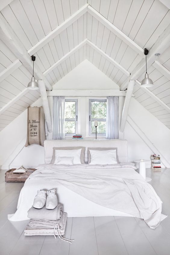 a white attic bedroom with an upholstered bed and white bedding, grey curtains, industrial lamps and a suitcase