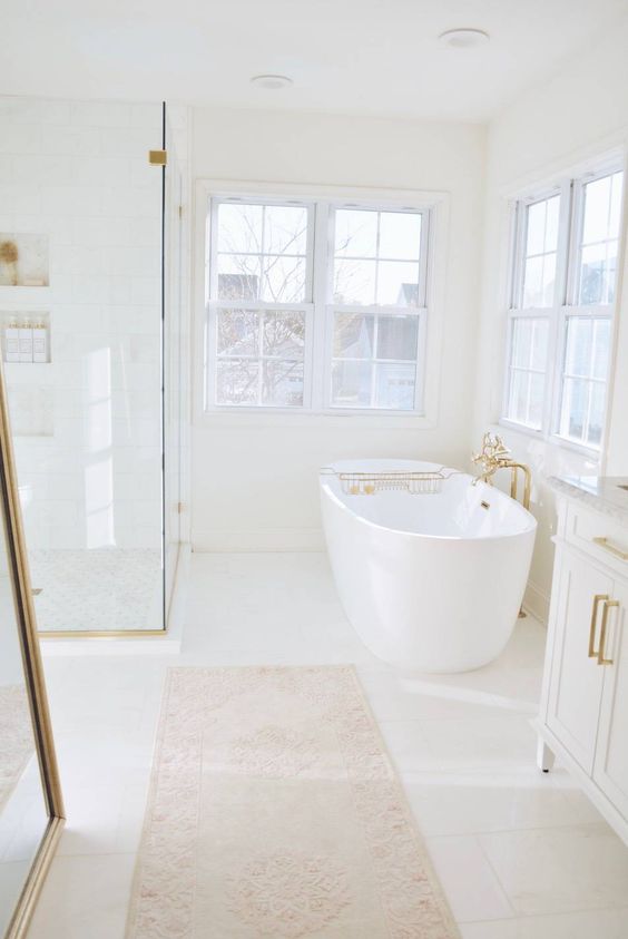 a white bathroom with large windows, a shower space enclosed in glass, an oval tub, a printed rug and gold fixtures