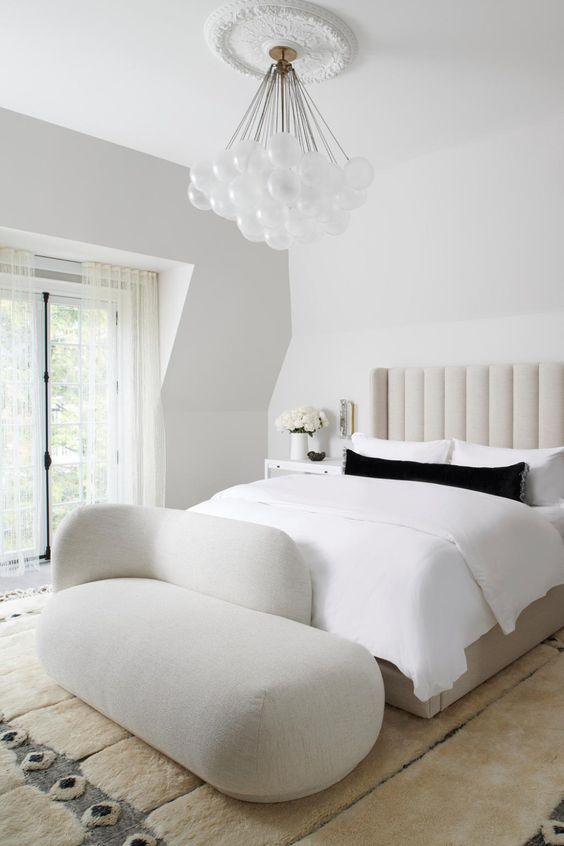 a white bedroom with a creamy upholstered bed, neutral bedding, a creamy daybed, a chandelier of bubbles