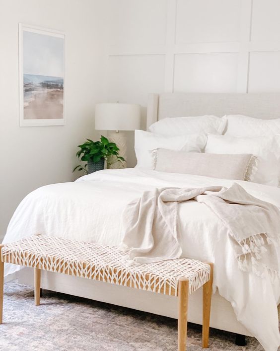 a white bedroom with a paneled accent wall, an upholstered bed with neutral bedding, a woven bench and an artwork