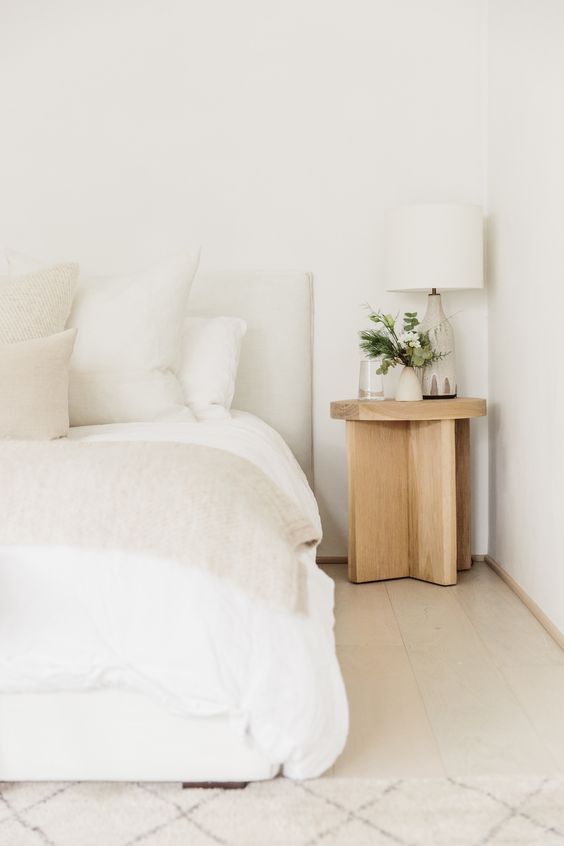 a white bedroom with an upholstered bed and neutral bedding, a stained nightstand and a white table lamp