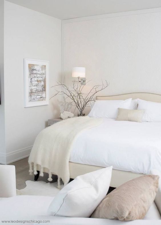 a white bedroom with an upholstered bed and white bedding, a daybed with neutral pillows, some art and branches