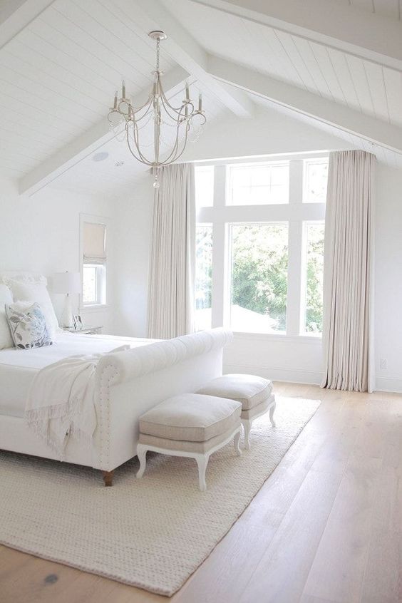 a white bedroom with an upholstered bed and white bedding, elegant neutral poufs and a vintage chandelier