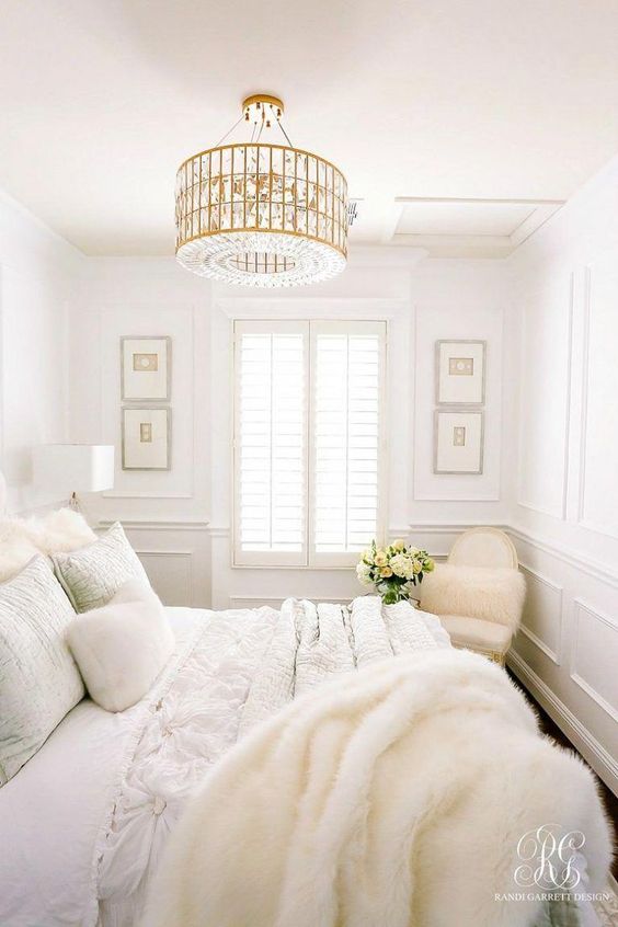 a white bedroom with paneled walls, a bed with neutral bedding, faux fur, a white chair and a chic gold chandelier