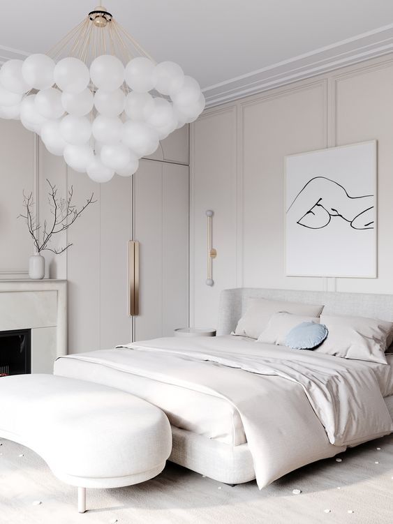 a white bedroom with paneling on the walls, a fireplace, an upholstered bed and a bench, a bubble chandelier