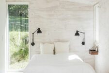 a white bedroom with plywood walls, a bed with white bedding, a couple of black sconces and jute poufs