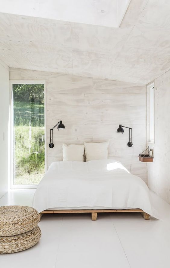 a white bedroom with plywood walls, a bed with white bedding, a couple of black sconces and jute poufs
