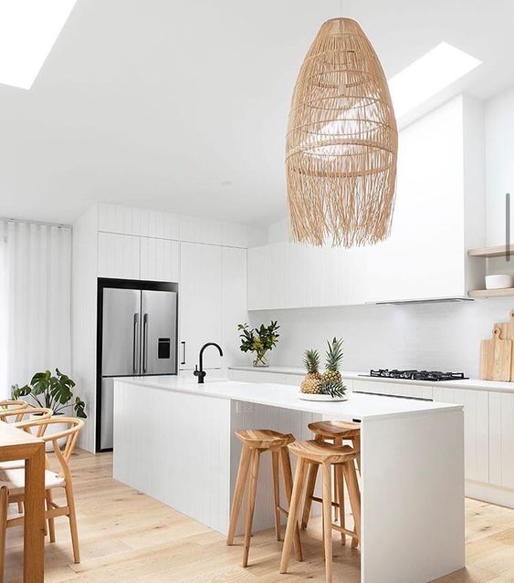 a white tropical kitchen with sleek cabinets, a white backsplash and countertops, stained stools and a woven pendant lamp