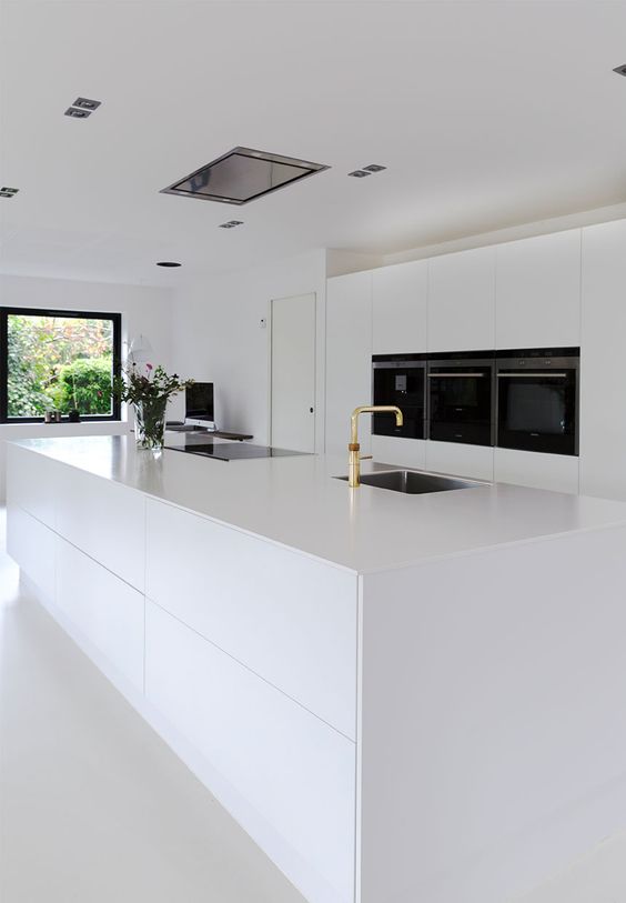 a white ultra-minimalist kitchen with sleek cabinets, an oversized kitchen island and built-in appliances plus gold fixtures
