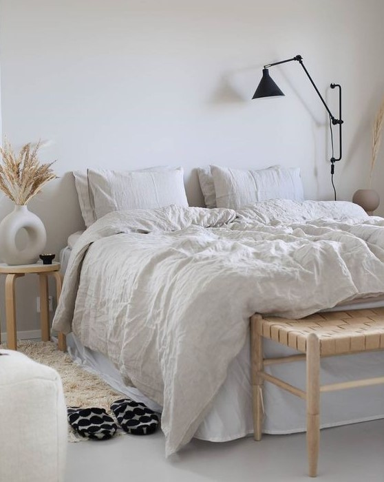 an airy neutral Scandi bedroom with modern furniture, a woven bench, neutral bedding, a fluffy rug, black sconces and grasses