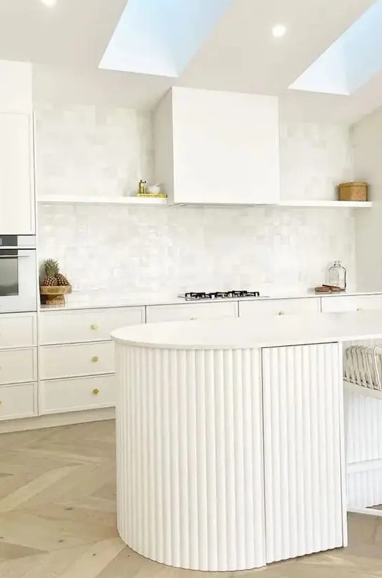 an airy white kitchen with white cabinets and an open shelf, a curved fluted kitchen island and skylights over it