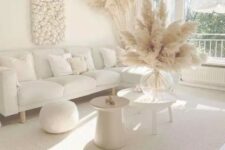 an all-white boho living room with a sectional, a pouf, a couple of side tables, pampas grass and a cool wall art