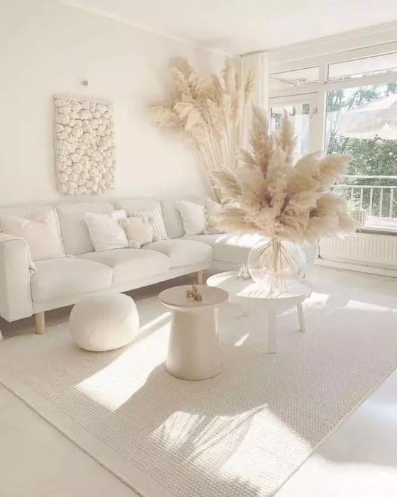 an all white boho living room with a sectional, a pouf, a couple of side tables, pampas grass and a cool wall art