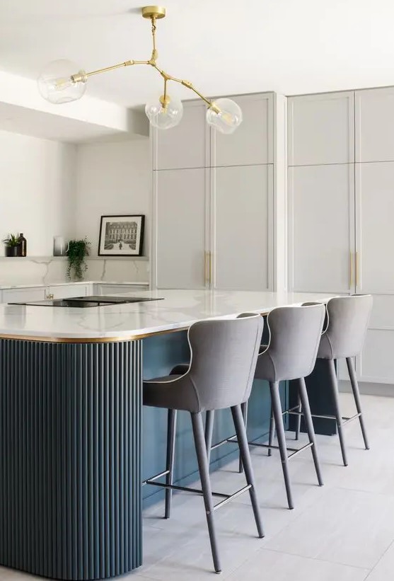 an exquisite dove grey kitchen with shaker cabinets, white marble countertops and a backsplash, a navy fluted kitchen island with a cooking and sitting zone