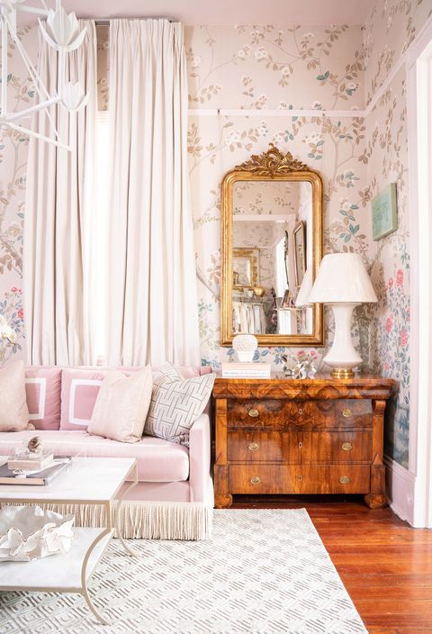 a catchy vintage living room with neutral floral wallpaper, a pink sofa with neutral pillows, a stained dresser, a mirror in a gold frame