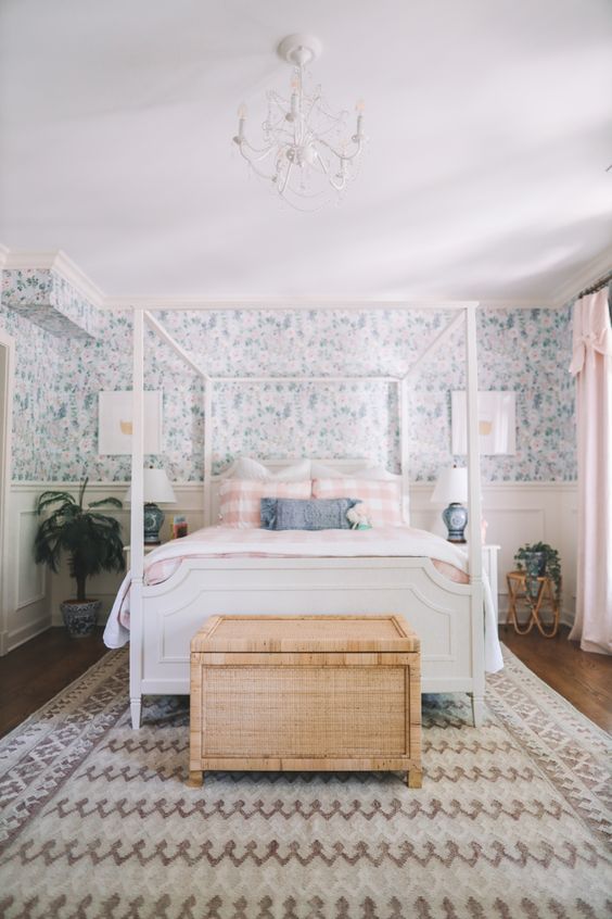 a cottage bedroom with blue floral wallpaper, a white bd with printed bedding, a woven chest, potted plants and a chandelier