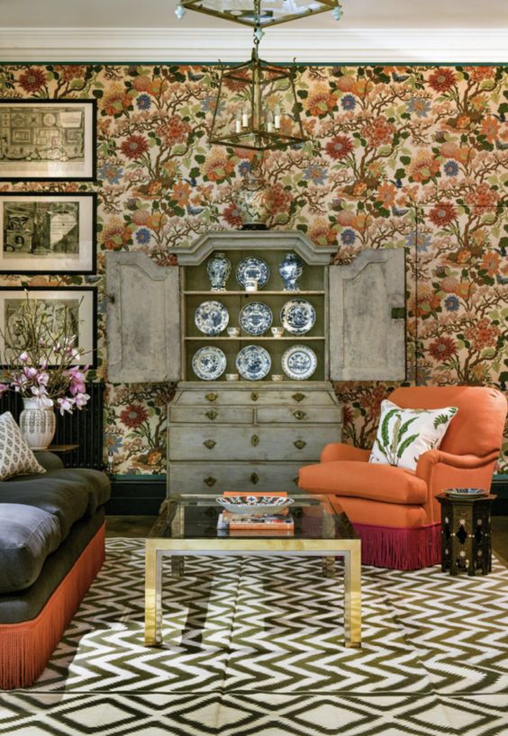 a maximalist cottagecore living room with bright floral wallpaper, a dark sofa and an orange chair, a black coffee table, a shabby chic buffet