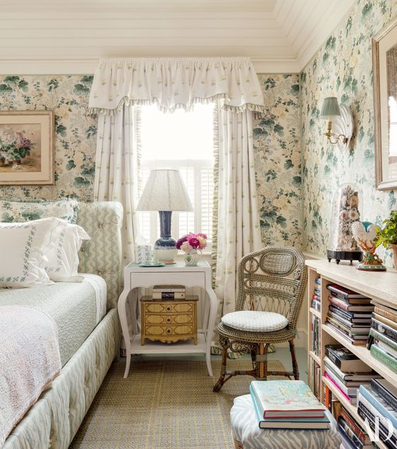 a cottage bedroom with floral wallpaper, a neutral bed with light green bedding, a white nightstand and a bookshelf