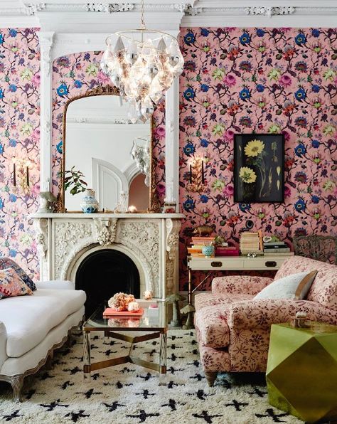a maximalist living room with pink floral wallpaper, an ornated fireplace, a white and a pink floral sofa, a printed rug and a chic chandelier