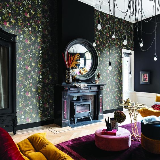 a moody living room with dark floral wallpaper, a black hearth, jewel-tone seating furniture and pendant bulbs