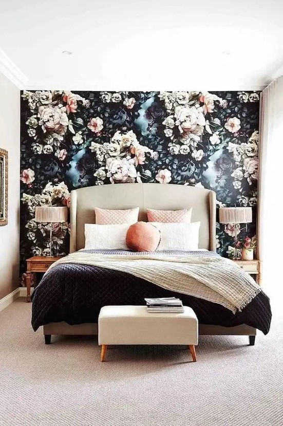a modern refined bedroom with a black and blush floral accent wall, a neutral upholstered bed, contrasting bedding, an upholstered stool