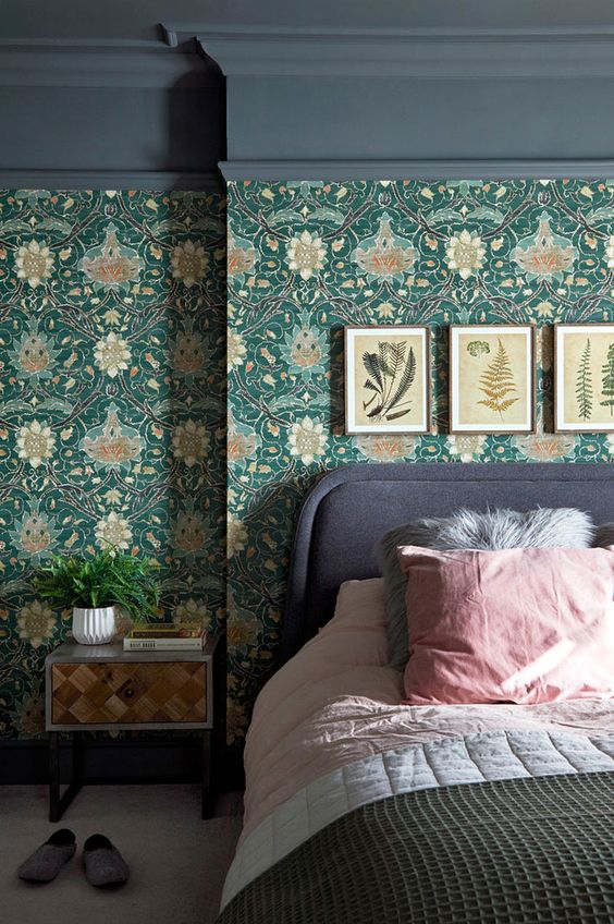 a moody bedroom with green floral wallpaper, a grey upholstered bed with pink bedding, a small nightstannd and a gallery wall