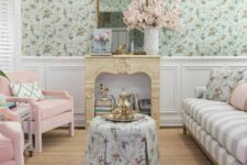 13 a pastel living room with green floral wallpaper, a striped sofa with pillows, pink chairs and a faux fireplace filled with books
