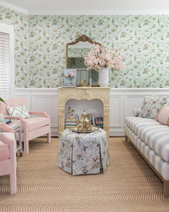 a pastel living room with green floral wallpaper, a striped sofa with pillows, pink chairs and a faux fireplace filled with books