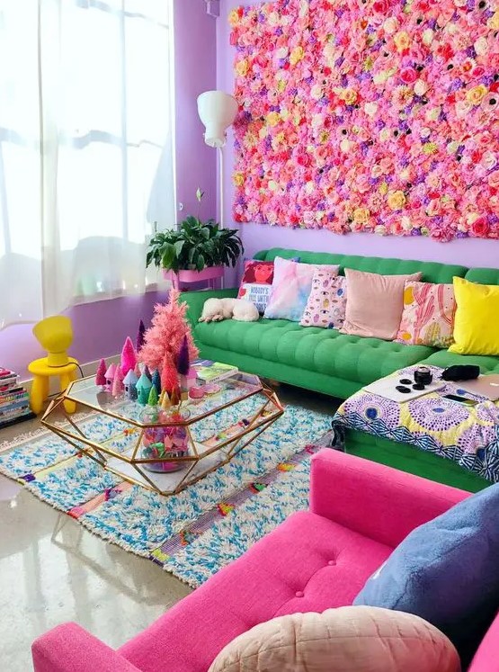 a super colorful maximalist living room with a bright faux flower wall, a green sofa, a hot pink chair, colorful pillows, a diamond-shaped glass table