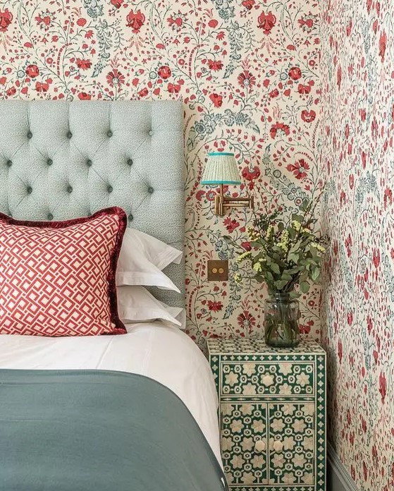 an eye-catchy bedroom with bold blue and red floral wallpaper, a mint upholstered bed with red printed and green bedding, a green and white floral nightstand