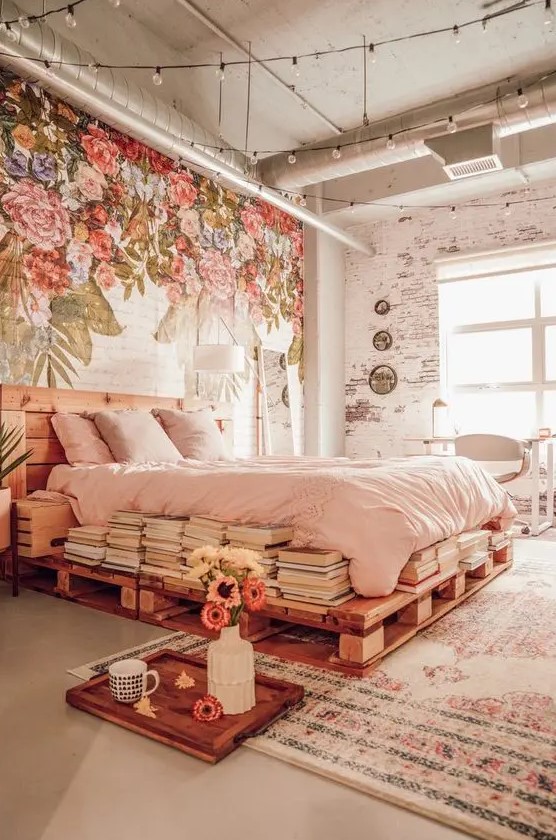 an eclectic bedroom with a painted floral wall, a pallet bed with blush bedding, stacks of books and bulbs over the space