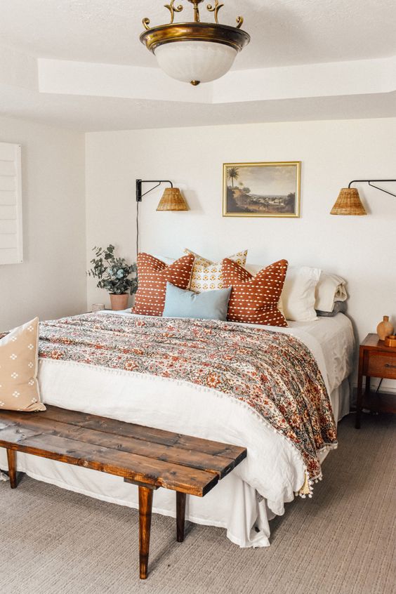 a pretty and welcoming bedroom with a bed and floral and white bedding, a stained bench, stained nightstands and a vintage chandelier