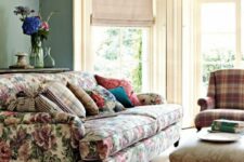 28 a vintage living room with green walls, a floral sofa and floral pillows, a neutral ottoman and a plaid chair plus stacks of books