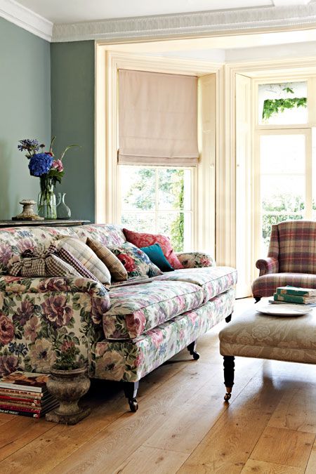 a vintage living room with green walls, a floral sofa and floral pillows, a neutral ottoman and a plaid chair plus stacks of books