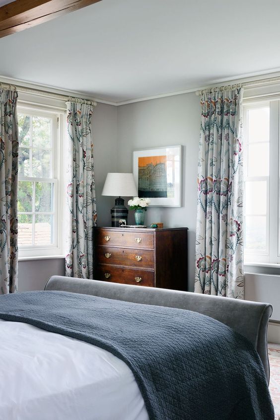 a neutral bedroom with light grey walls, a grey upholstered bed with neutral bedding, floral curtains, a stained dresser and a table lamp