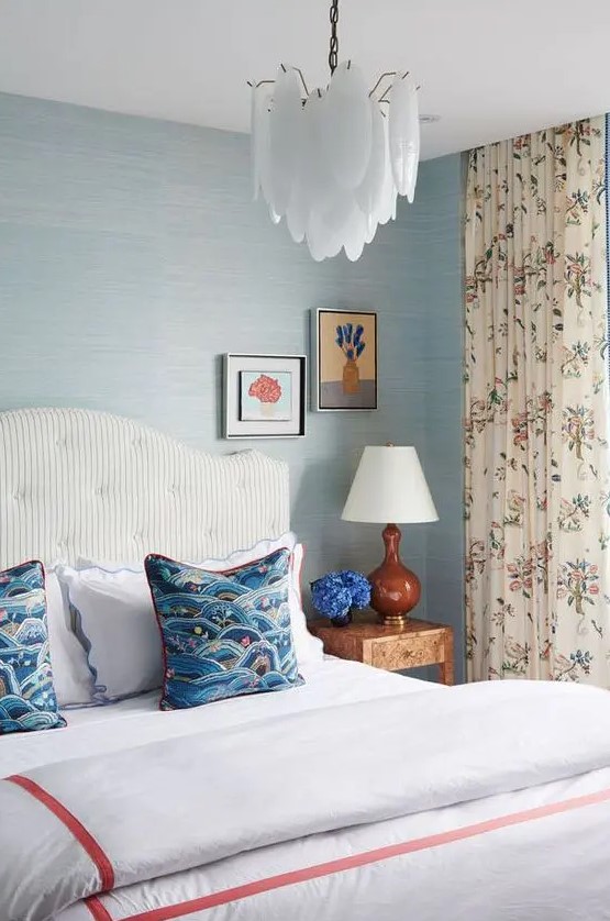 a breezy bedroom with blue wallpaper walls, a white striped bed with blue bedding, floral curtains and a catchy scallop chandelier