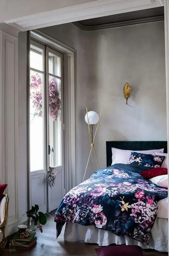 a chic bedroom with concrete walls, a black bed with pink, navy and black floral bedding, chic gilded lamps and a stack of books