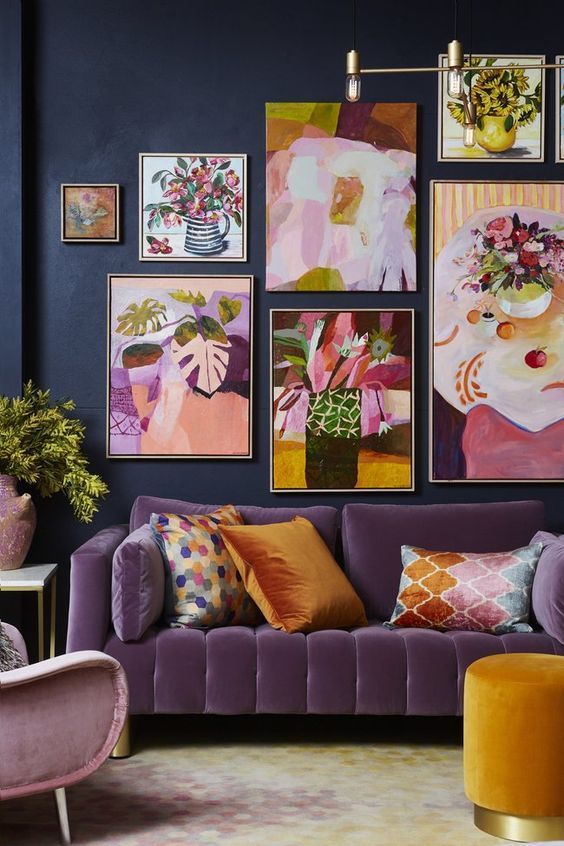 a moody living room with navy walls, a purple sofa and a pink chair, a rust pouf and a super bold floral gallery wall