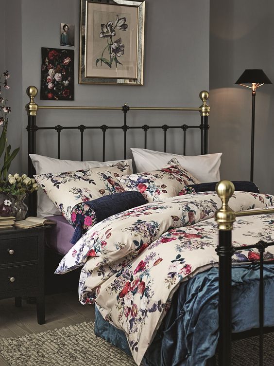 a moody bedroom with grey walls, a black forged bed with bold floral bedding, a black nightstands and a black floor lamp